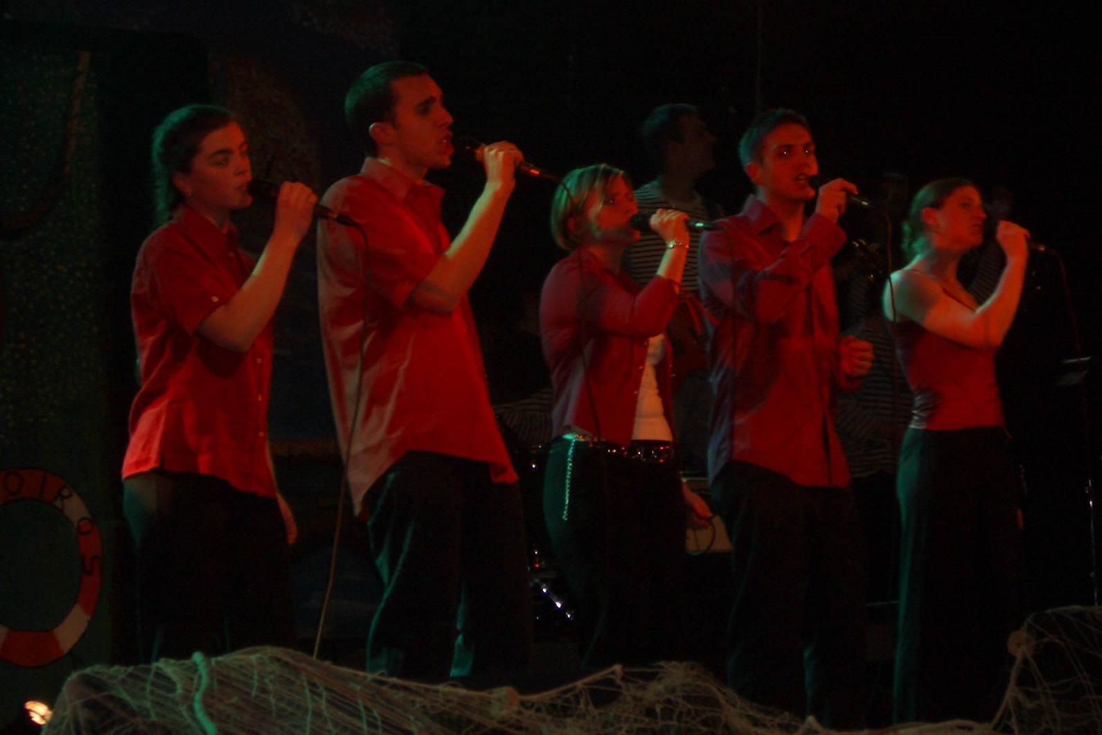 Photograph of five people on a stage (three women and two men), singing into microphones. They are dressed with red shirts and black pants. The second person from the left is Guillaume Paumier.
