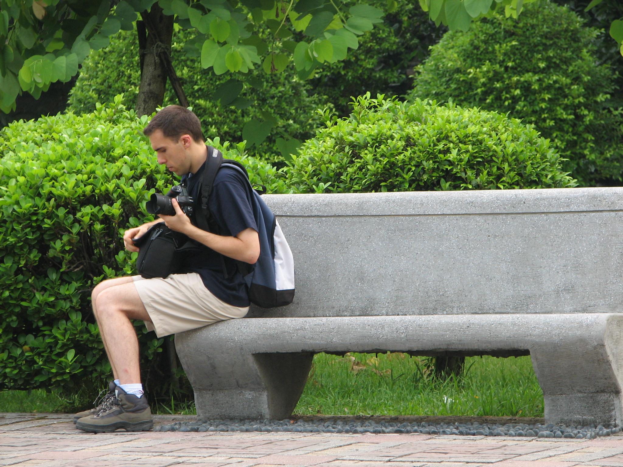 A picture of Guillaume Paumier sitting on a bench, changing a lens on a camera, on a background of green plants.