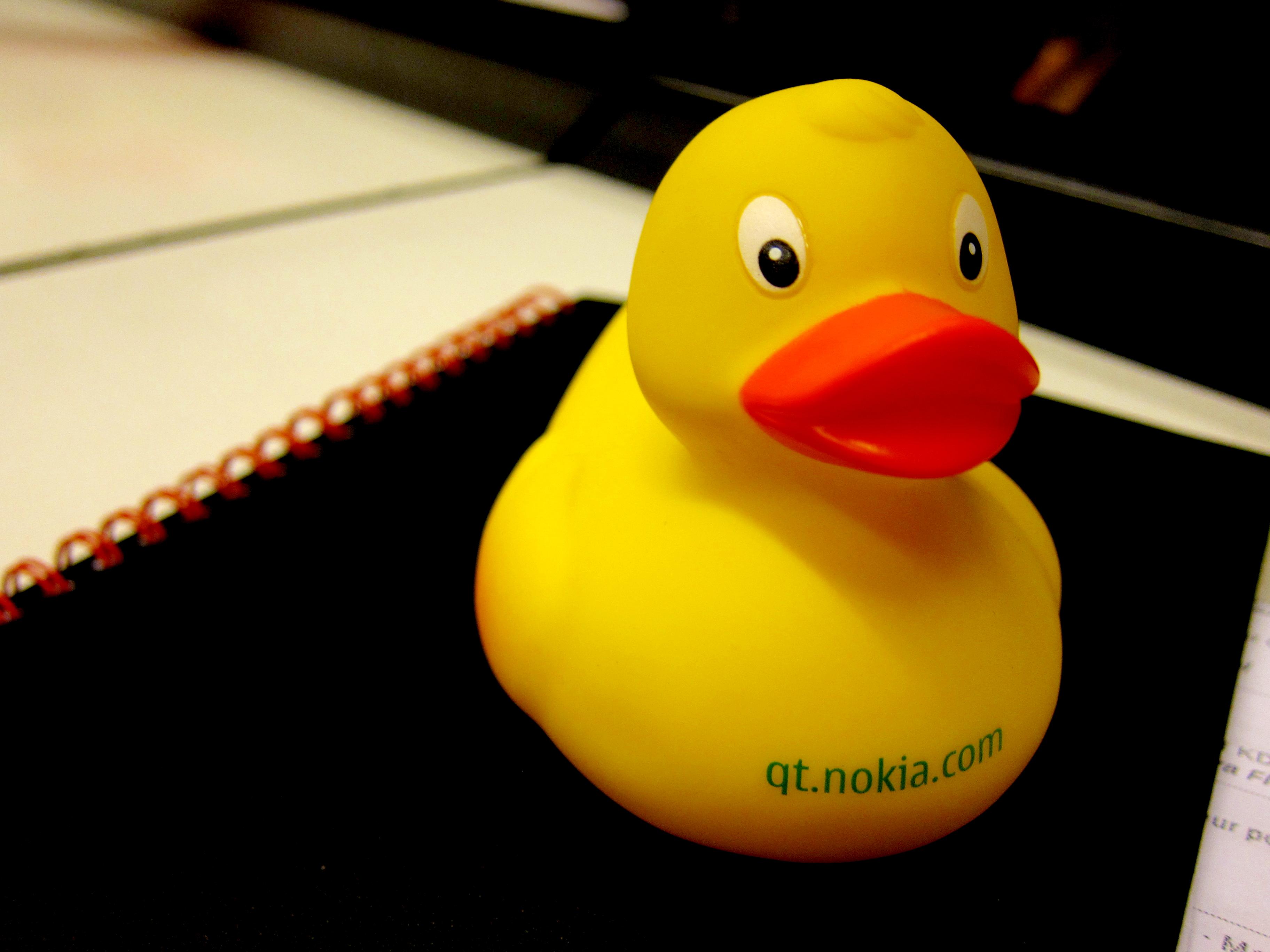 A bright yellow rubber duck with an orange beak sits atop a black notebook with a red spiral binding. The words “qt.nokia.com” are printed in green on the duck’s side.