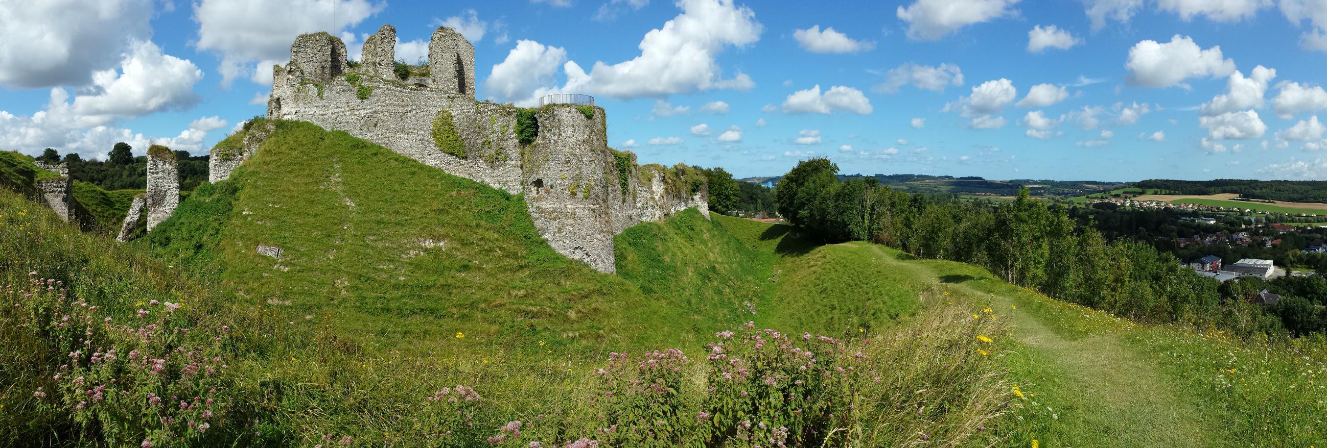 Panoramic view of an old stone castle overseeing a green landscape, under a cloudy blue sky.
