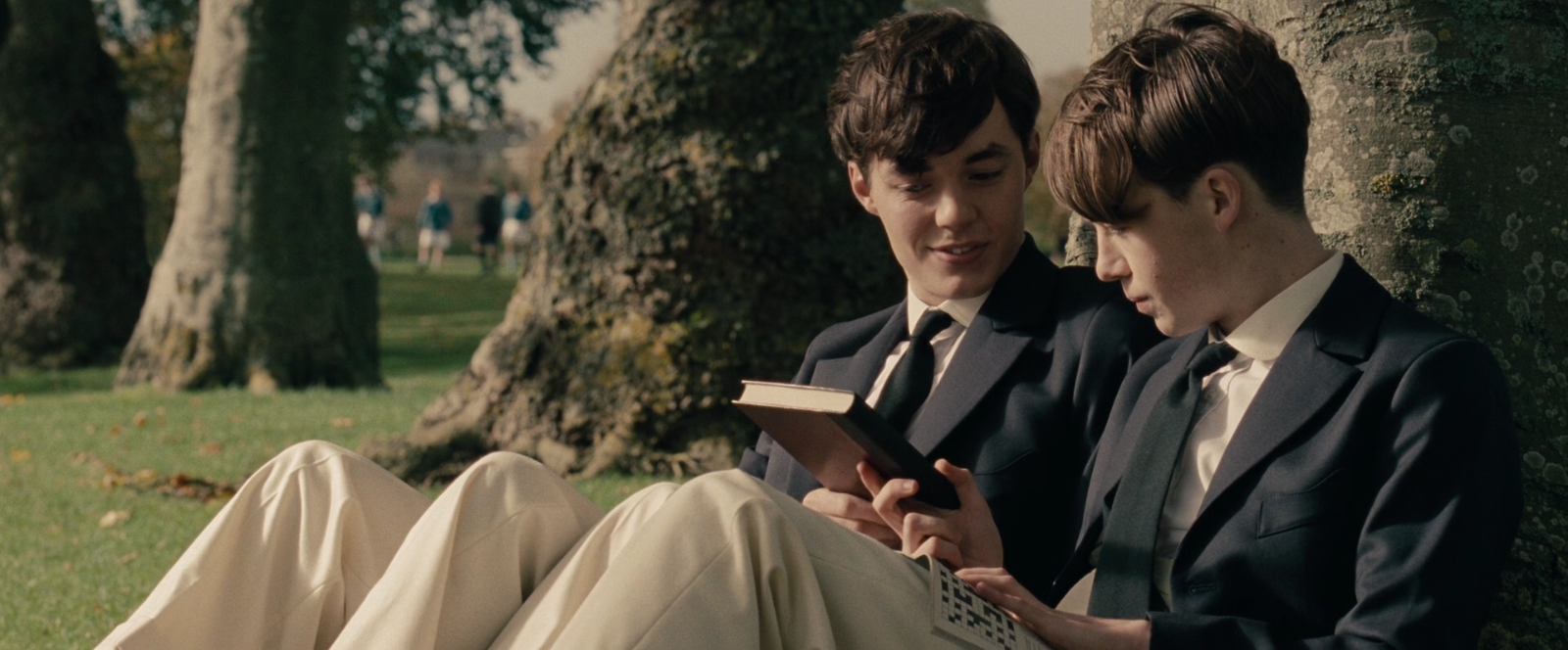 A screenshot of the movie The Imitation Game, showing a young Alan Turing (played by Alex Lawther) and his friend Christopher Morcom (portrayed by Jack Bannon). They are sitting against a tree, and Christopher is handing Alan a book.