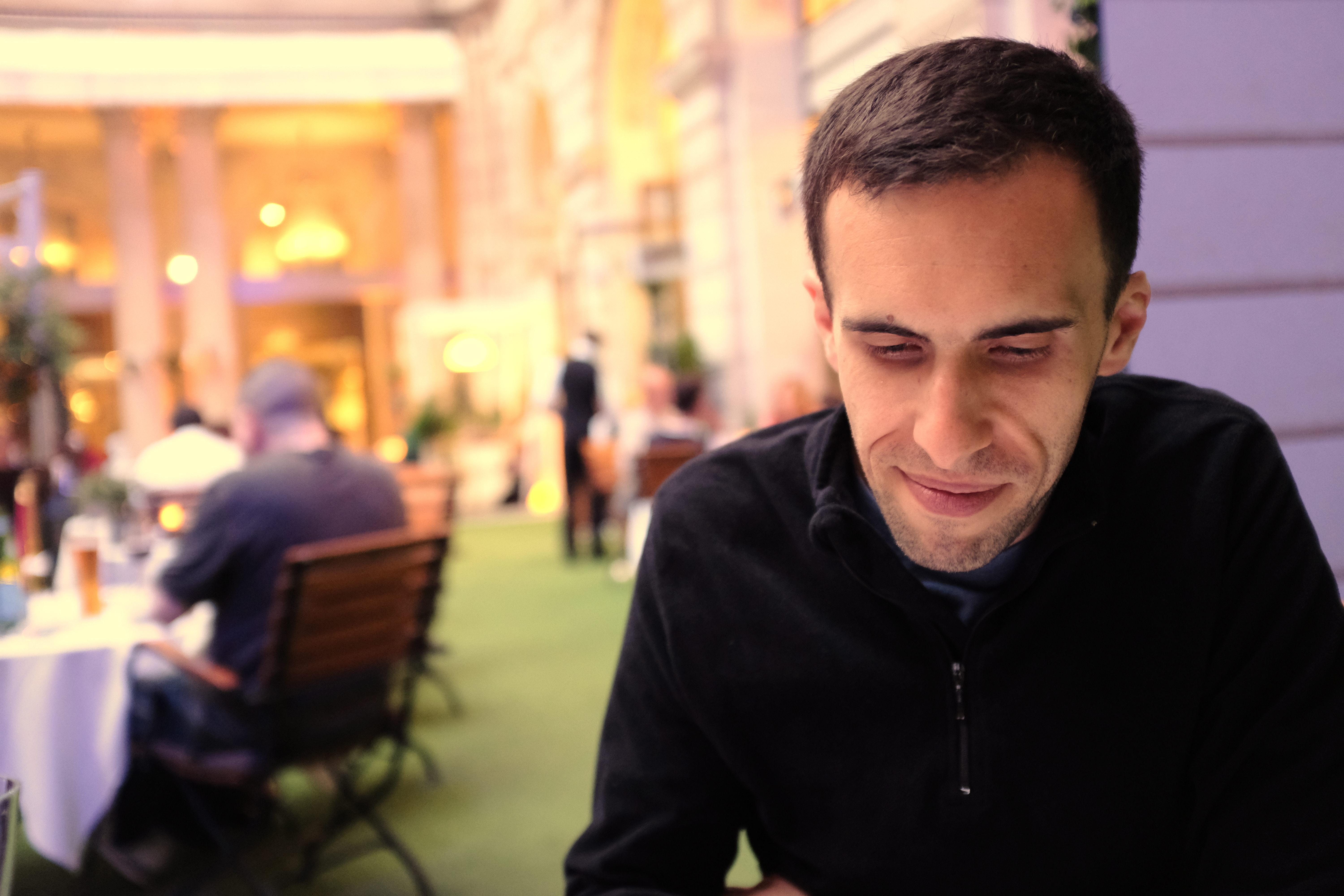 Photograph of Guillaume Paumier. He is a white man with short brown hair, slightly smiling, looking down, and wearing a black fleece. Behind him is the blurry background of a Parisian restaurant, showing warm hues.