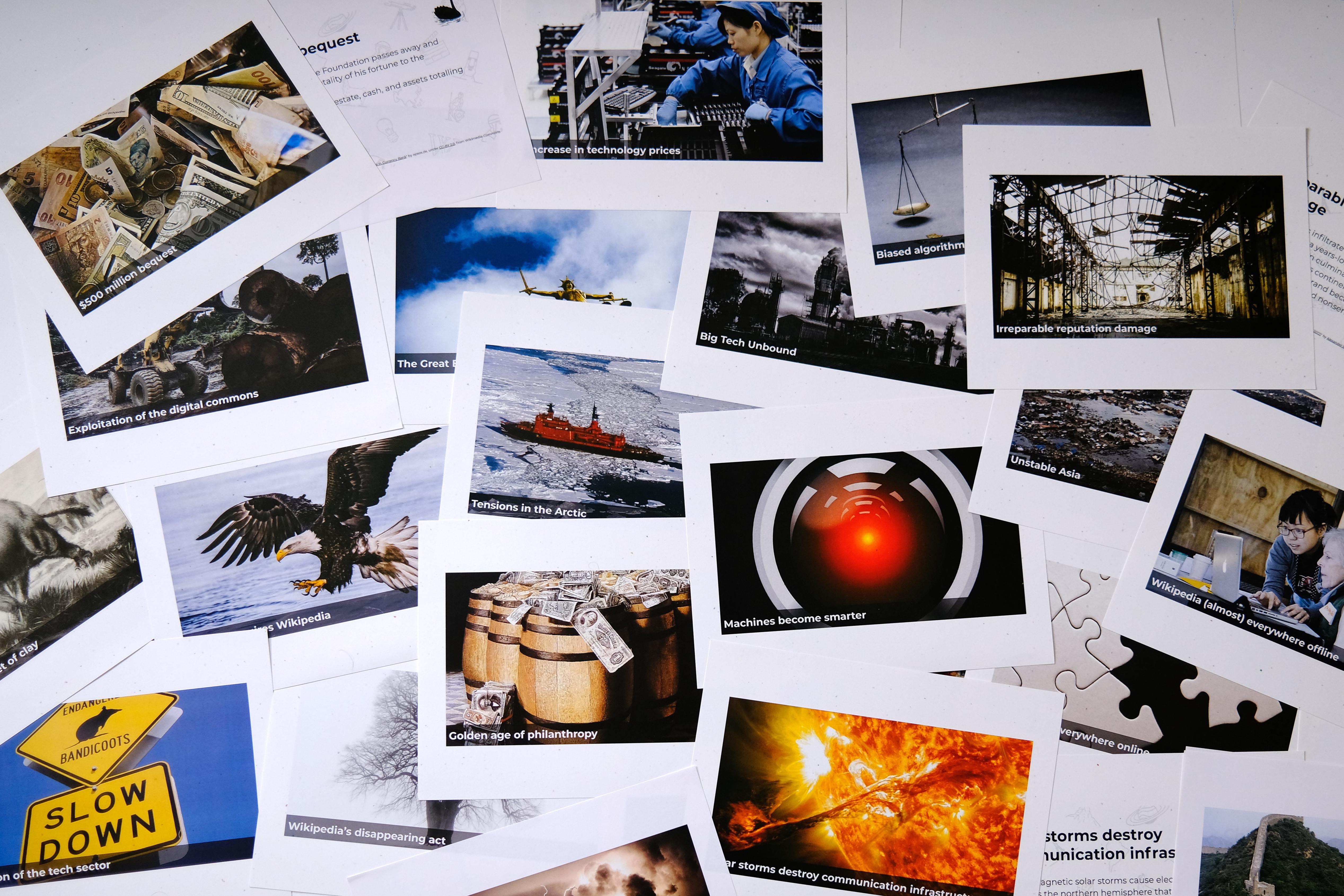 Photograph of two dozen scenario cards printed on large sheets of thick paper, showing various images with titles, and messily arranged on a surface.