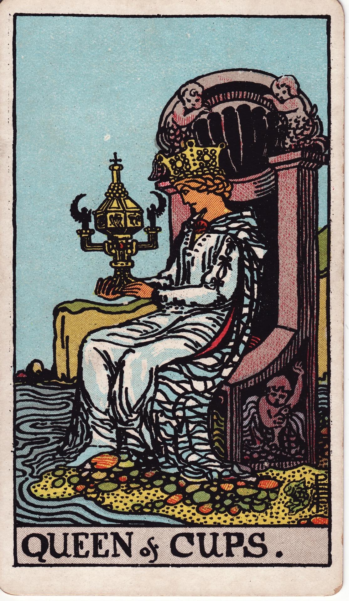 The Queen of Cups card from the Rider–Waite tarot deck. A crowned woman in flowing robes sits on a stone throne by the sea, gazing thoughtfully into an ornate, lidded chalice. The throne is adorned with carvings of sea nymphs, and her feet rest lightly on colorful stones at the water’s edge.